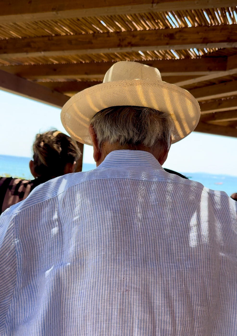 An older man in a straw hat and blue striped shirt sits under a pergola facing the sea, sunlight casting patterns; ocean in background.