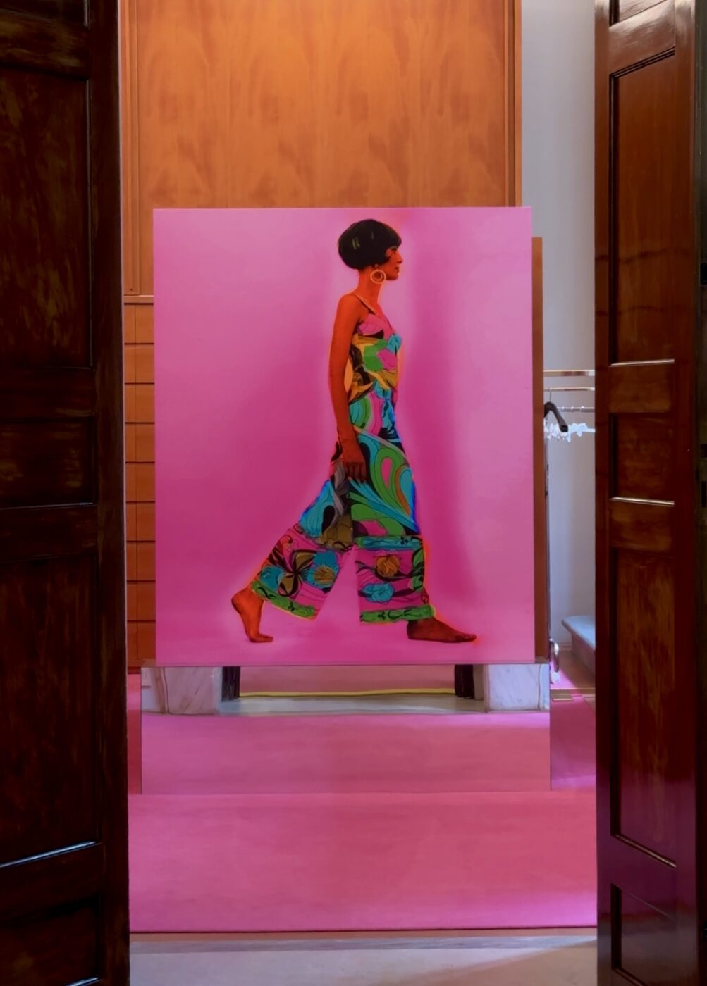 A vibrant painting of a woman in a patterned jumpsuit on a pink background hangs between two open doors in a modern room.