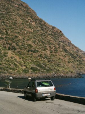 A small gray car sits by a coastal road near a calm blue sea, with green hills and rocky slopes under a clear sky in the background.
