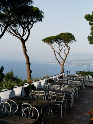 Terrazza esterna con tavoli, sedie, fiori rossi sul muro, vista sul mare, alti pini, montagne lontane sotto un cielo limpido.