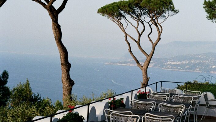 An outdoor terrace with empty metal chairs, tables, pine trees, and flower pots overlooks the sea under a clear blue sky.