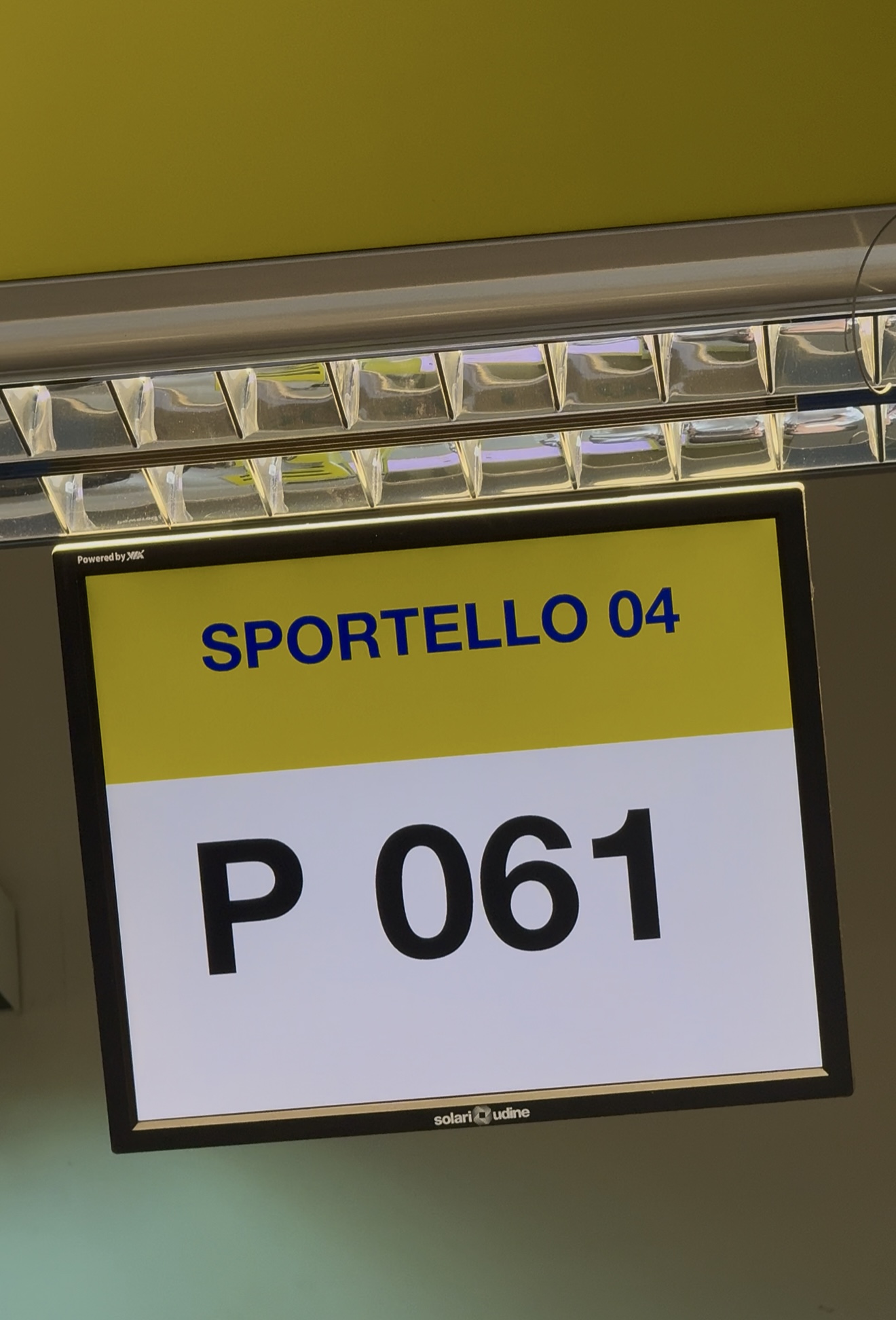 Un cartello digitale indica SPORTELLO 04 in blu su giallo, con P 061 in grande nero sotto, che indica un numero di coda o di sportello.