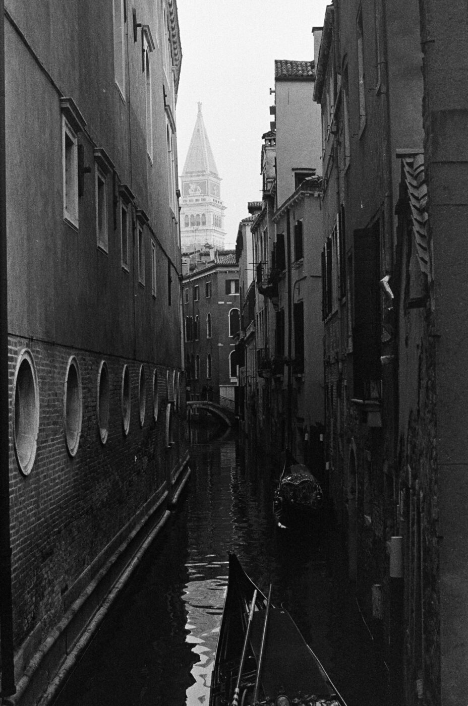 A black and white Venice canal scene with a gondola, tall buildings, and a distant bell tower above rooftops.