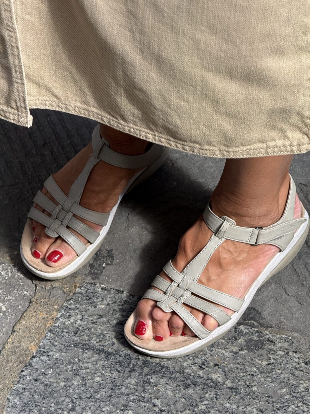 A person in beige sandals with red toenails stands on gray stone pavement, partially covered by a light tan skirt or pants.