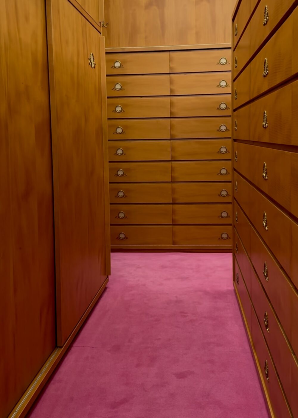 A narrow walk-in closet with wood cabinets, gold handles, and drawers lining both sides, plus a bright pink carpeted floor.