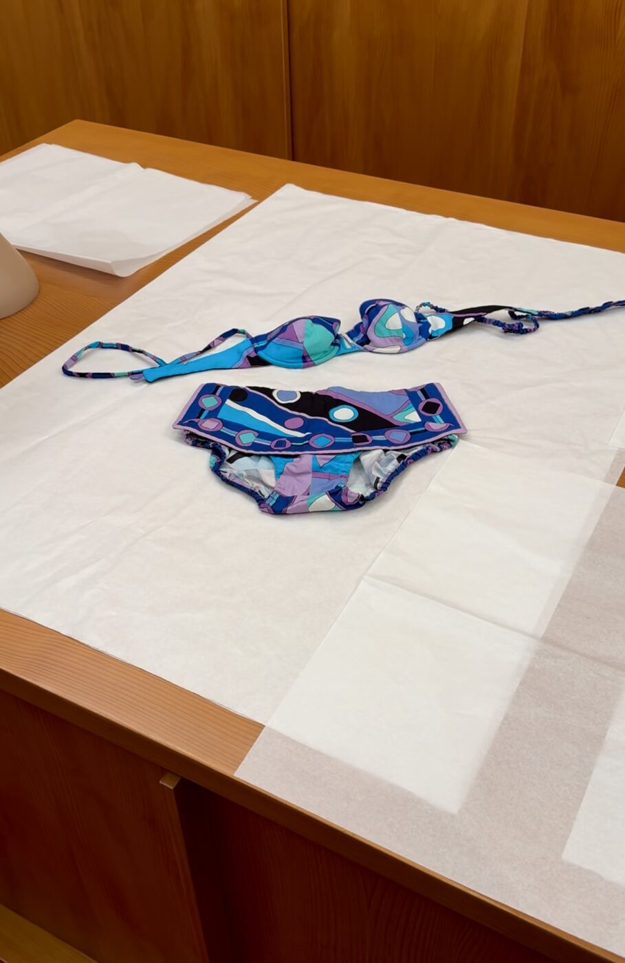 A bikini with blue, purple, and teal geometric patterns and white circles is spread on white paper atop a wooden table.