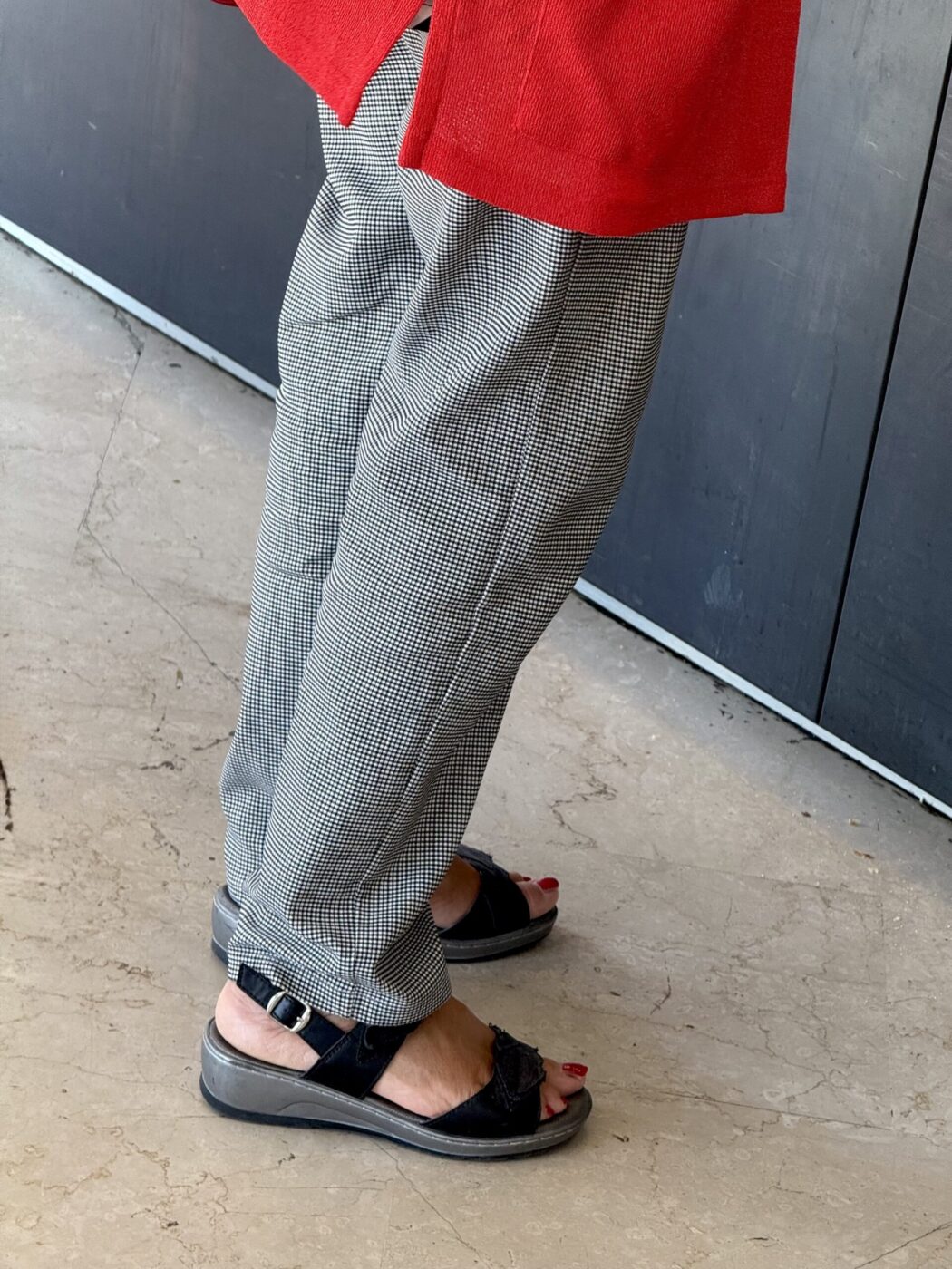 A person in gray checkered pants, red top, and black sandals with painted toenails stands on beige marble by a dark wall.