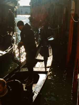 A gondolier stands on a gondola in a sunlit Venetian canal, with reflections on the water and more gondolas beside old buildings.
