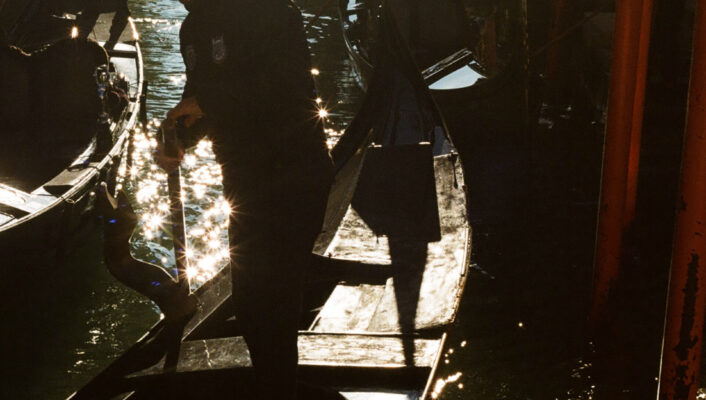 A gondolier stands on a gondola in a sunlit Venetian canal, with reflections on the water and more gondolas beside old buildings.