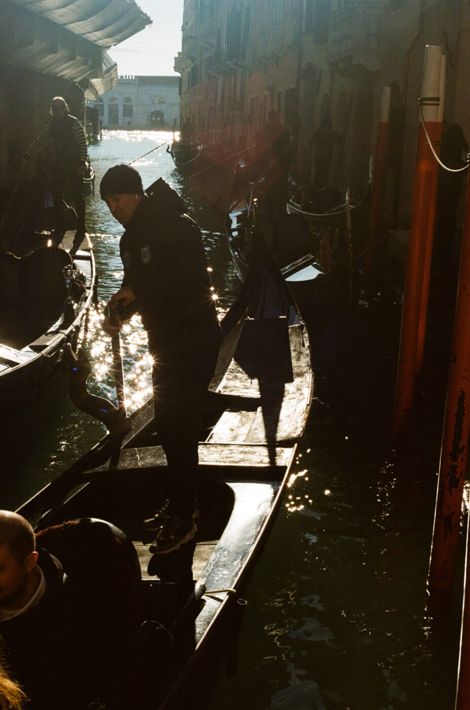 A gondolier stands on a gondola in a sunlit Venetian canal, with reflections on the water and more gondolas beside old buildings.