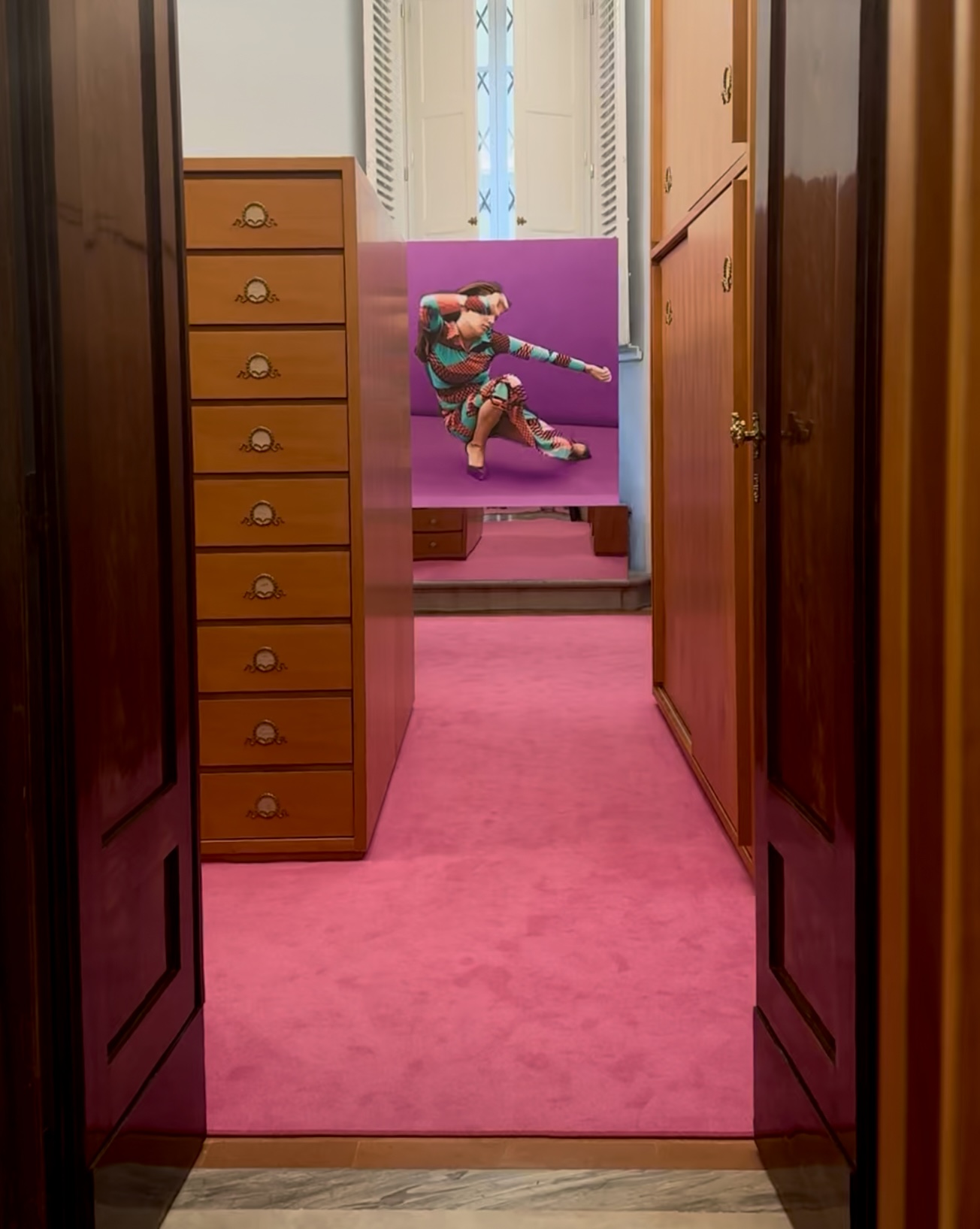 View through wooden doors into a room with pink carpet, wood cabinets, and bold art of a figure in vibrant clothes on purple.