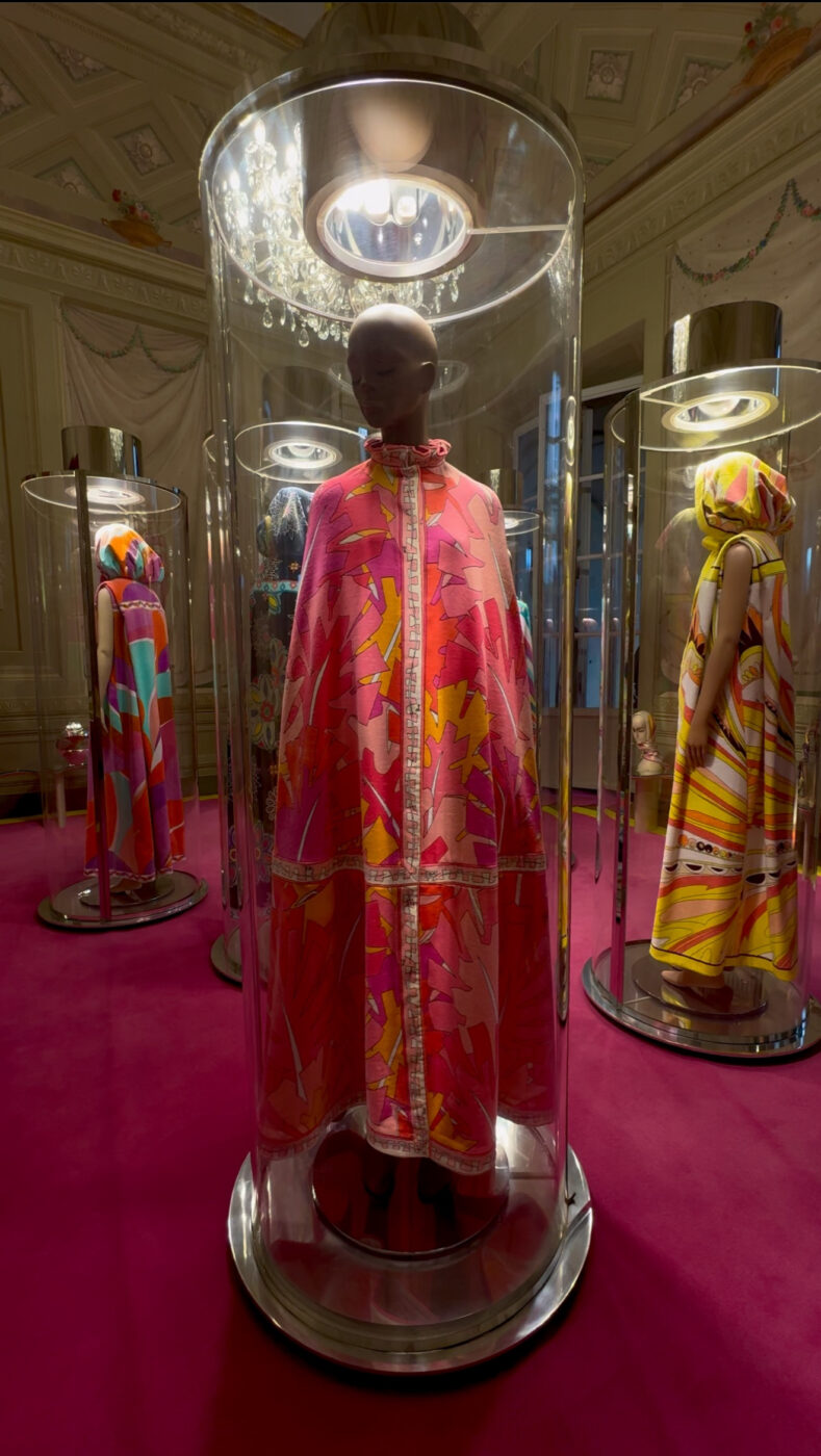 Three mannequins in bright patterned dresses and capes stand in glass cases on magenta carpet; center wears a pink-orange floral cape.