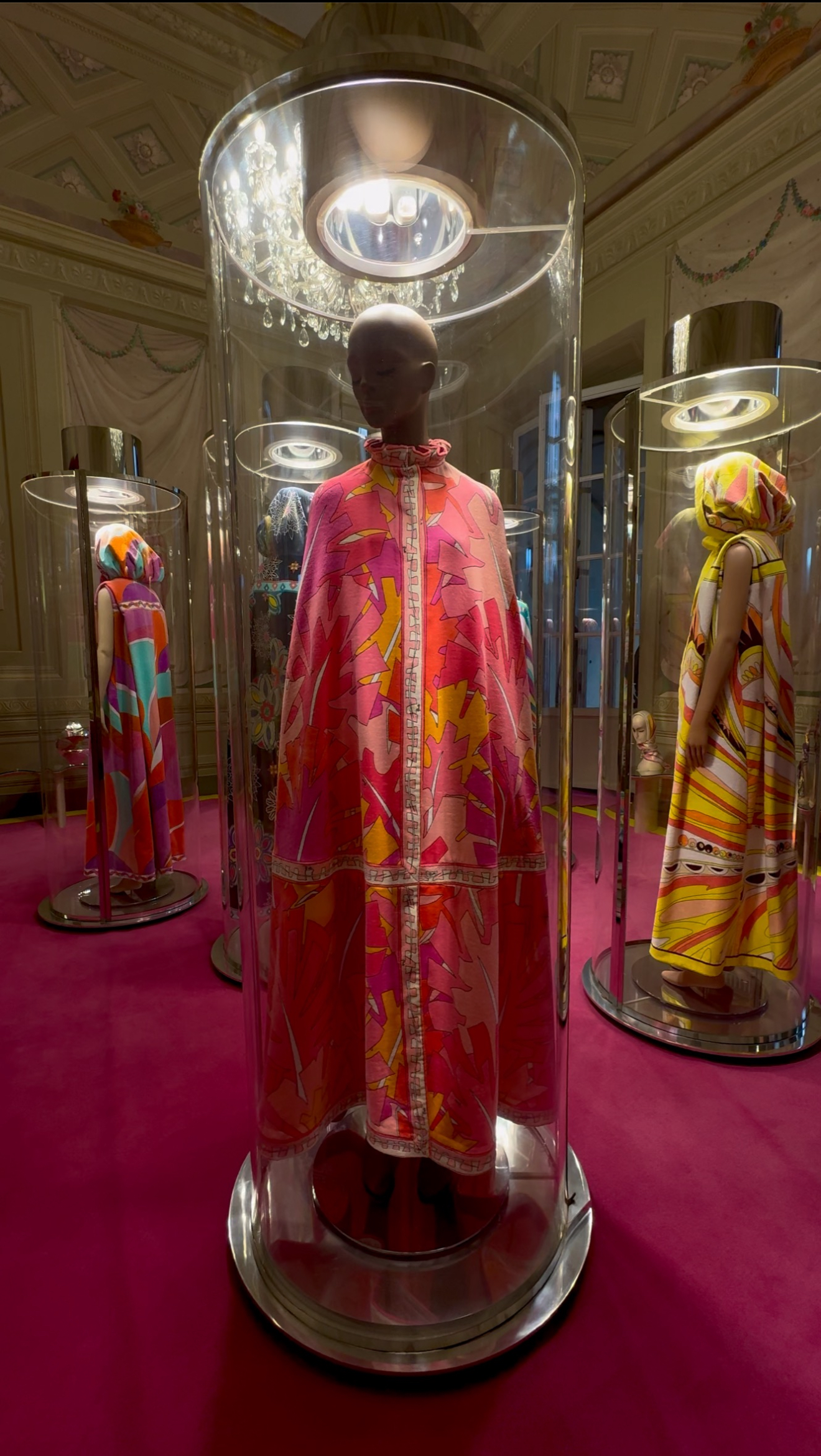 Three mannequins in bright patterned dresses and capes stand in glass cases on magenta carpet; center wears a pink-orange floral cape.