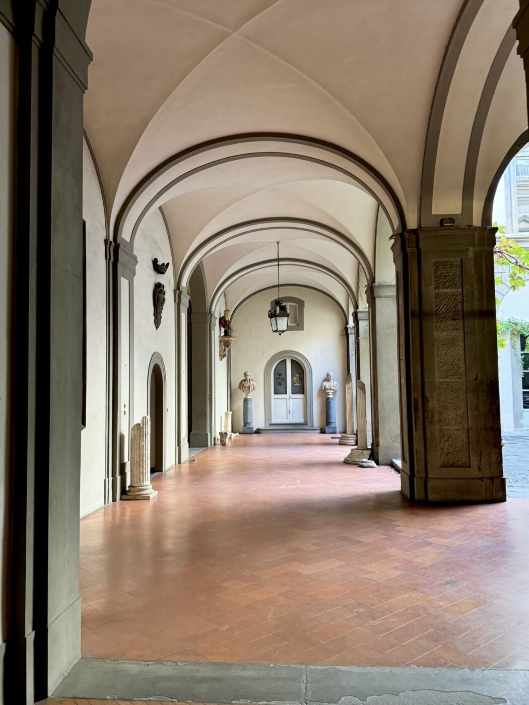 A covered walkway with arched ceilings, pale walls, terracotta tiles, classical statues, and sunlight from an open courtyard at right.