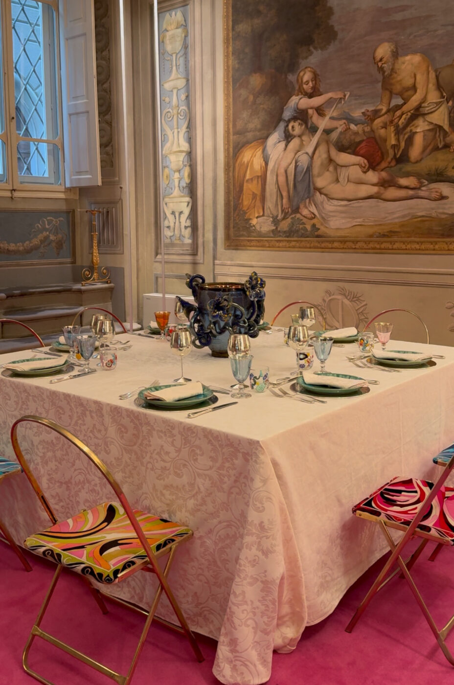A square table for six with a pale cloth, vibrant patterned chairs, and blue centerpiece stands in an ornate room with classical art.
