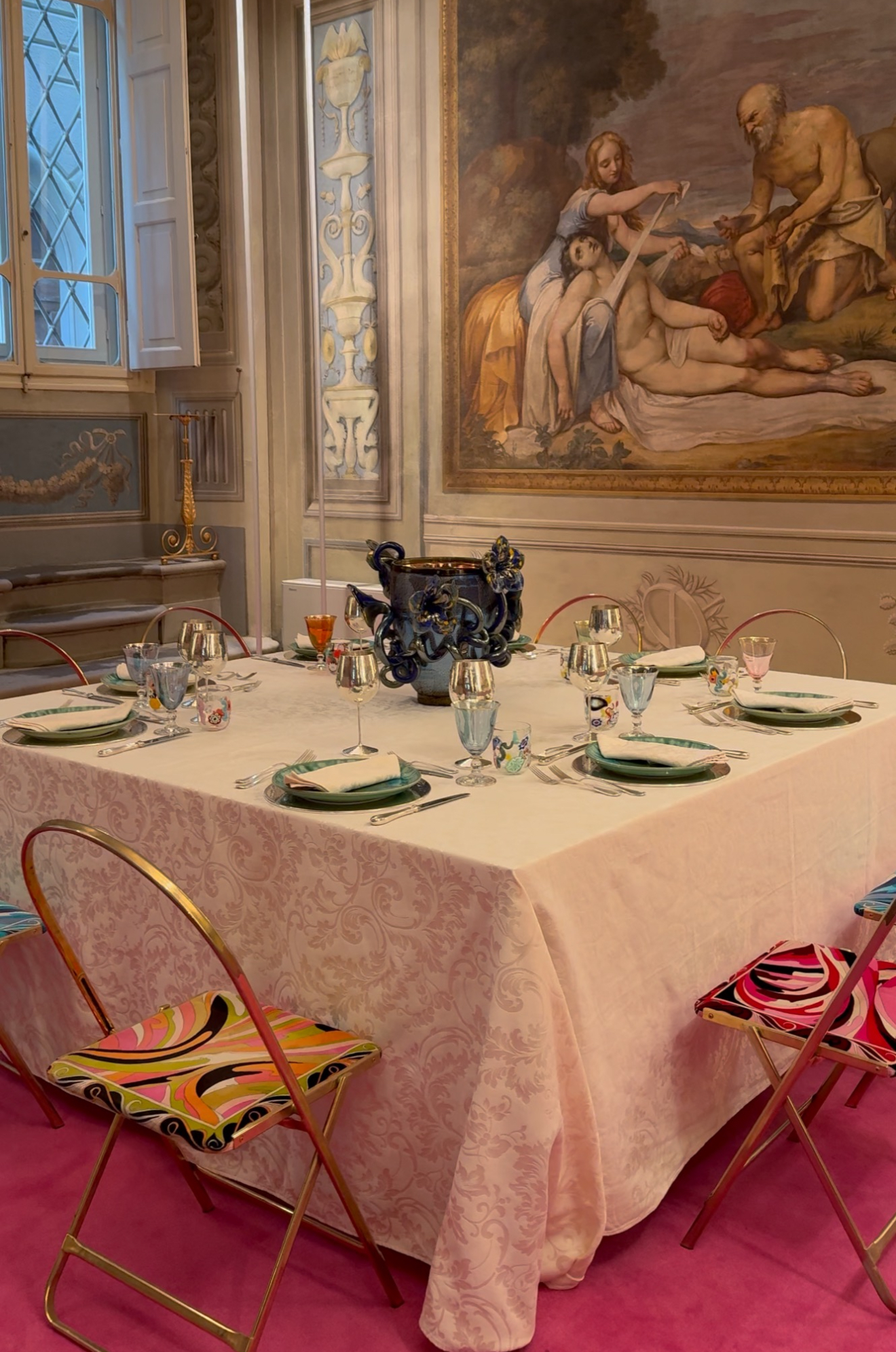 A square table for six with a pale cloth, vibrant patterned chairs, and blue centerpiece stands in an ornate room with classical art.