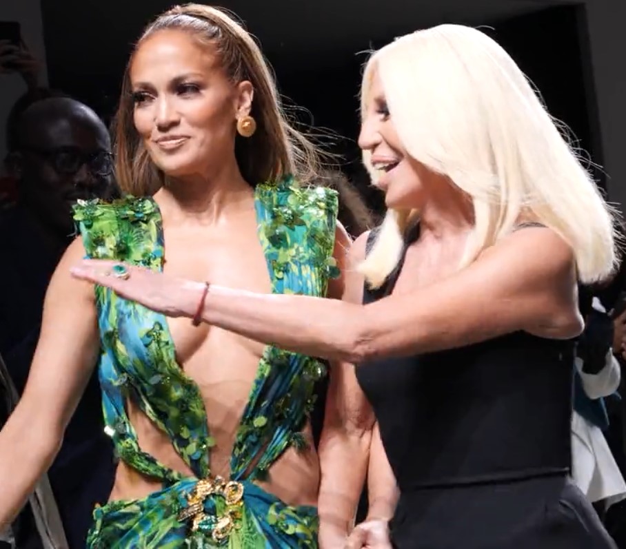 J.Lo and Donatella Versace on a runway: one in a green leaf-print dress, the other in black with long blonde hair; both are smiling.
