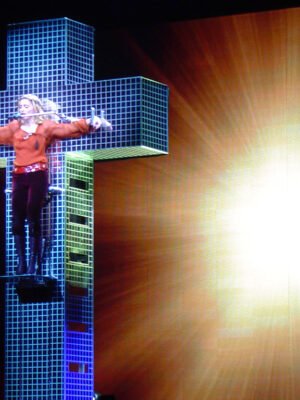 Un performer su una piattaforma illuminata a forma di croce con una griglia, con le braccia aperte, si trova davanti a un fondale luminoso giallo-arancio.