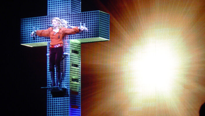 Madonna stands atop a glowing cross-shaped stage, arms spread, radiant beams and bright light illuminating the background.
