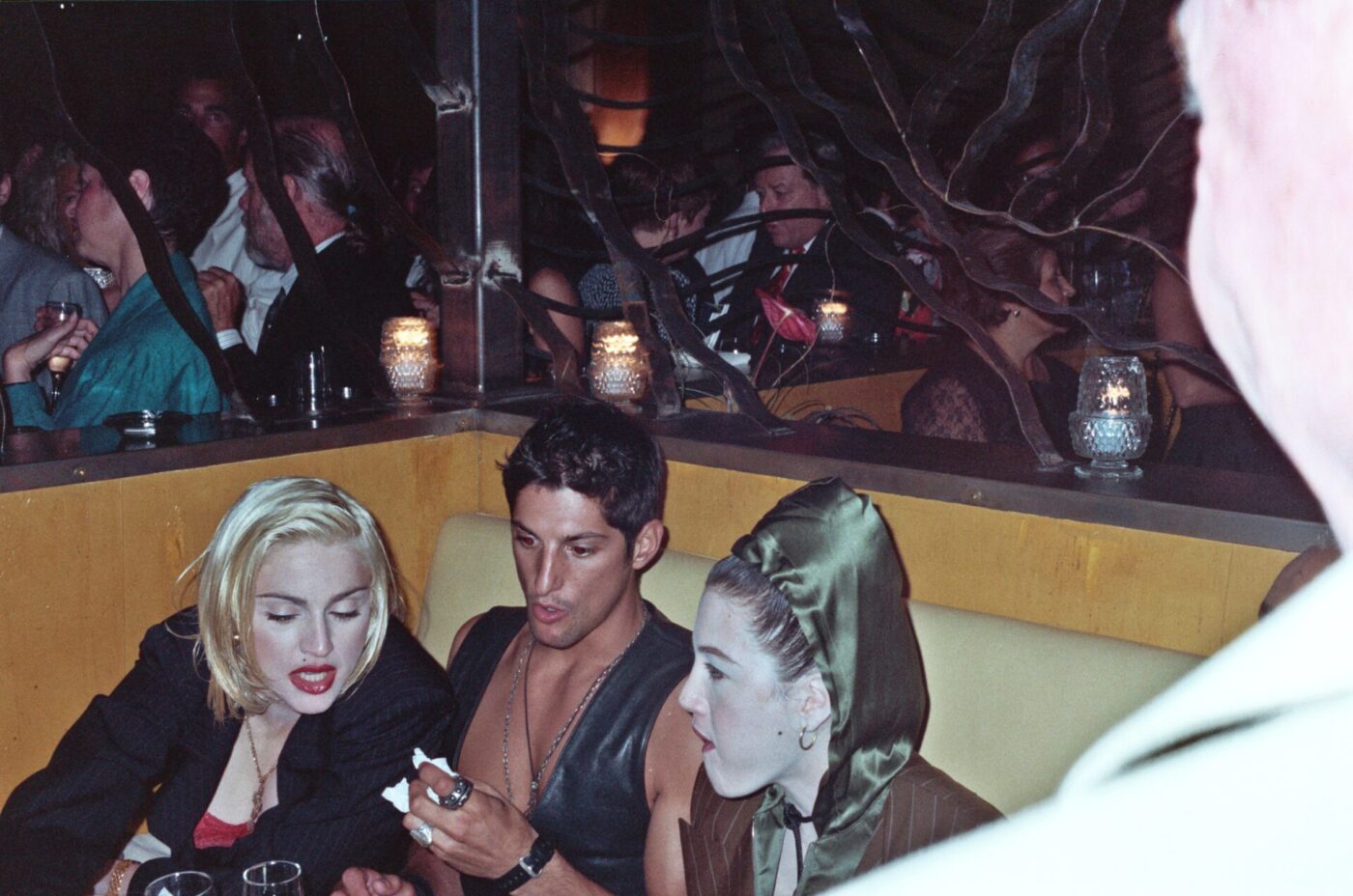 Three people sit close in a dim nightclub booth; two women, one with blonde hair, one in a green hood, and a man talk near candles.