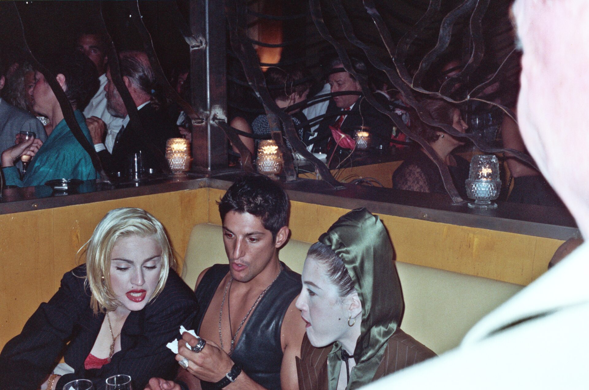 Three people sit close in a dim nightclub booth; two women, one with blonde hair, one in a green hood, and a man talk near candles.
