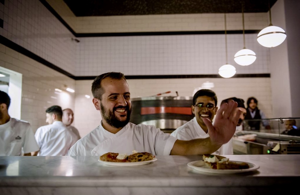 A smiling chef in white uniform grabs a plate on a marble counter; kitchen staff work behind under round lights in the restaurant.