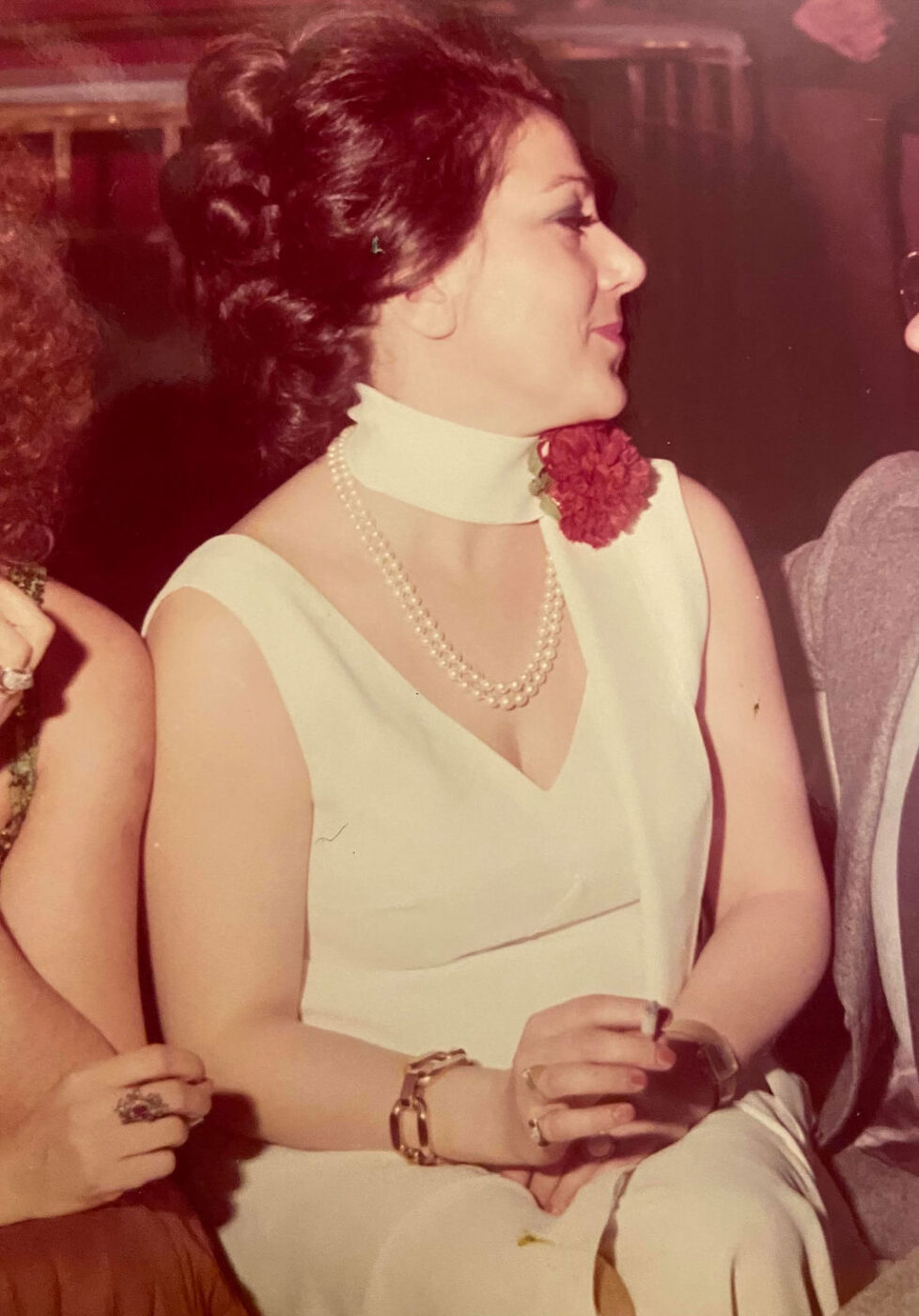 Una donna in abito bianco senza maniche con perle e un fiore rosso al collo sorride, guardando di lato con le mani in grembo.