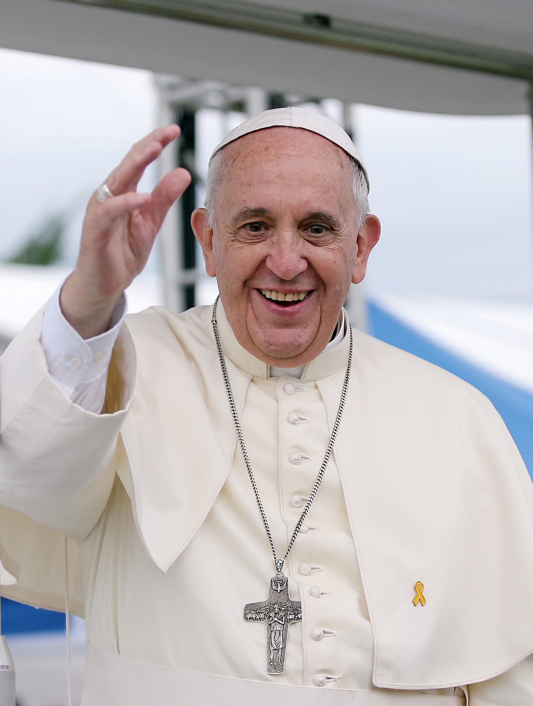 A smiling man in white papal robes and skullcap waves, cross necklace and yellow ribbon visible, outdoors under a white canopy.