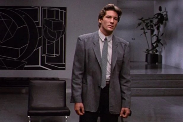 A man in a gray suit stands in a minimalist room with geometric art and a potted plant, looking ahead with a serious expression.