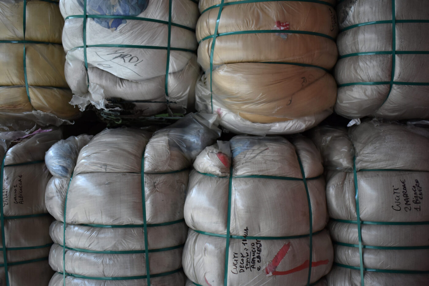 Tightly wrapped bales of fabric or clothing with green straps, stacked in rows; some have handwritten labels on the plastic wrapping.
