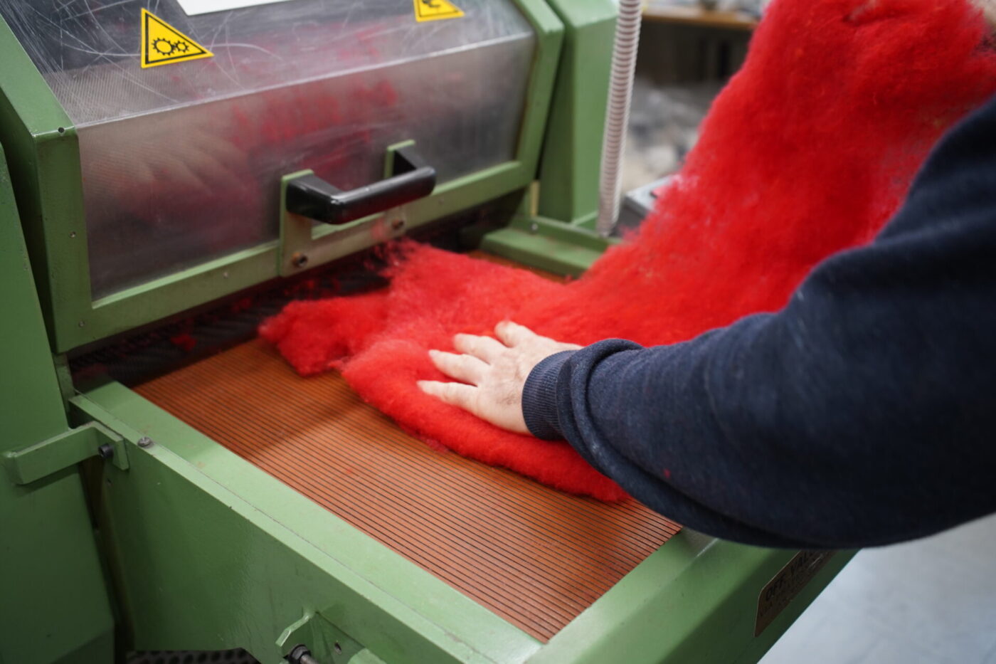 A person’s hand feeds vivid red wool into a green carding machine inside a textile factory.
