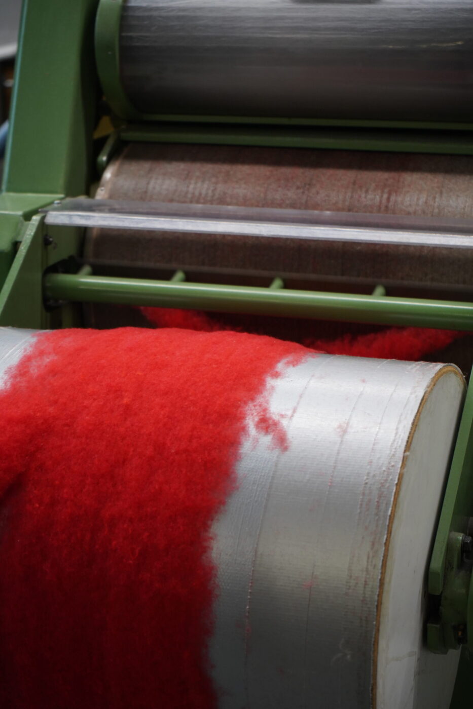 A close-up of a machine processing bright red wool as fibers are spread and smoothed over large cylindrical rollers.