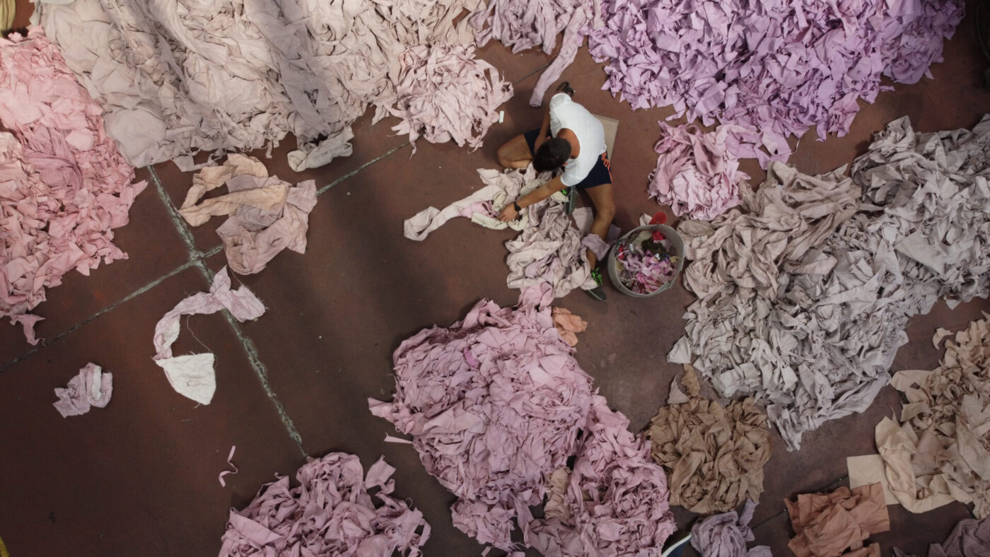 A person on a reddish floor sorts through large heaps of pink and purple fabric scraps, seen from above.