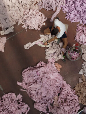 A person on a reddish floor sorts through large heaps of pink and purple fabric scraps, seen from above.