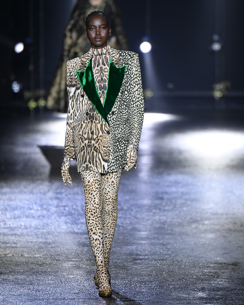 A model in a leopard print bodysuit and green velvet-lapel blazer struts a glossy black runway under dramatic lights.