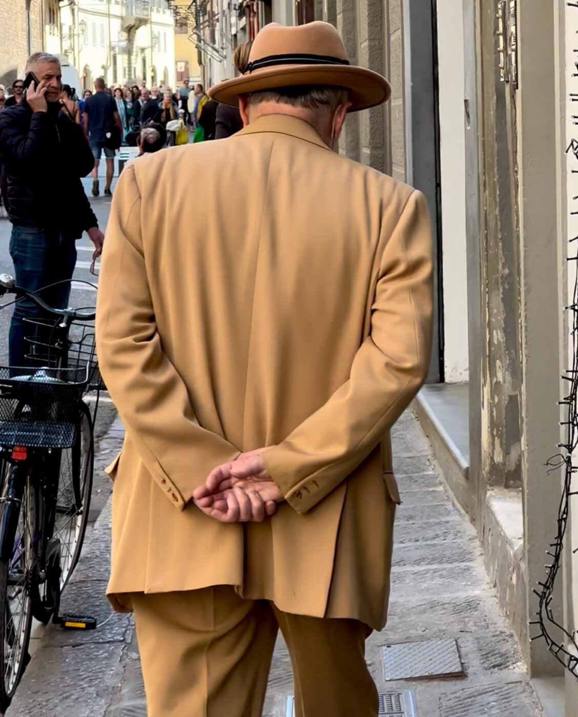 A man in a tan suit and hat strolls a narrow city street, hands behind his back; a bicycle and people appear in the background.
