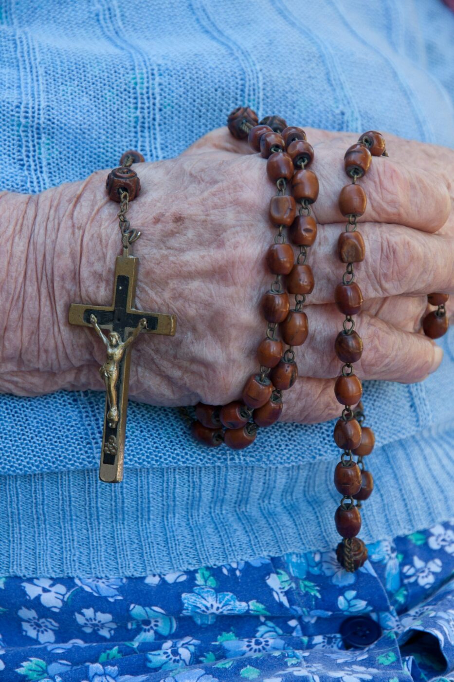 An elderly person’s hand holds a wooden rosary with crucifix on their lap, wearing a light blue sweater and floral blue pants.