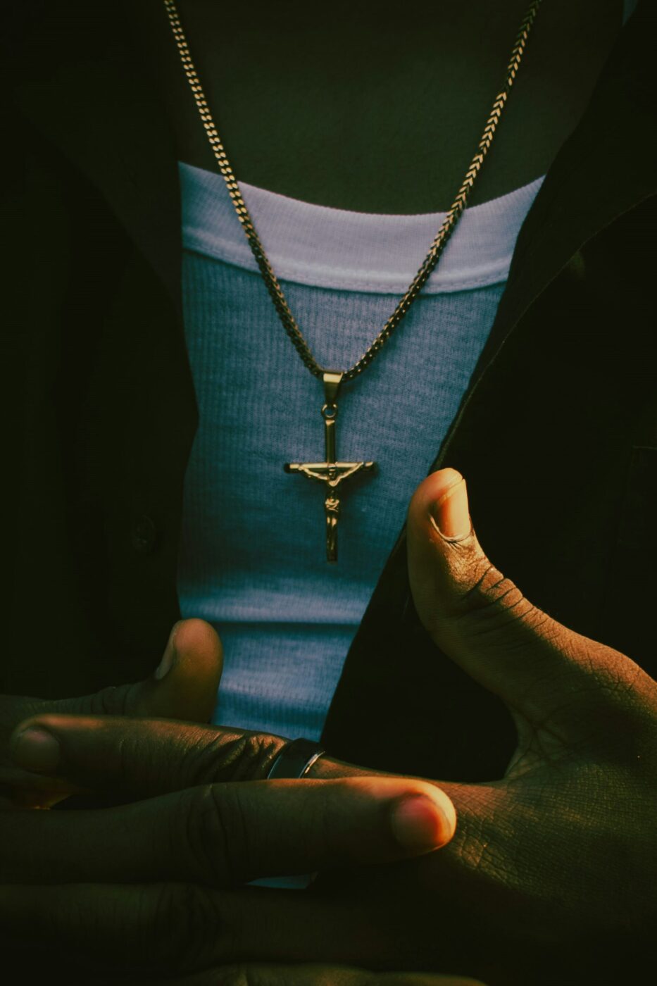 A person in a white tank top and dark shirt, gold crucifix necklace visible, hands clasped in front.