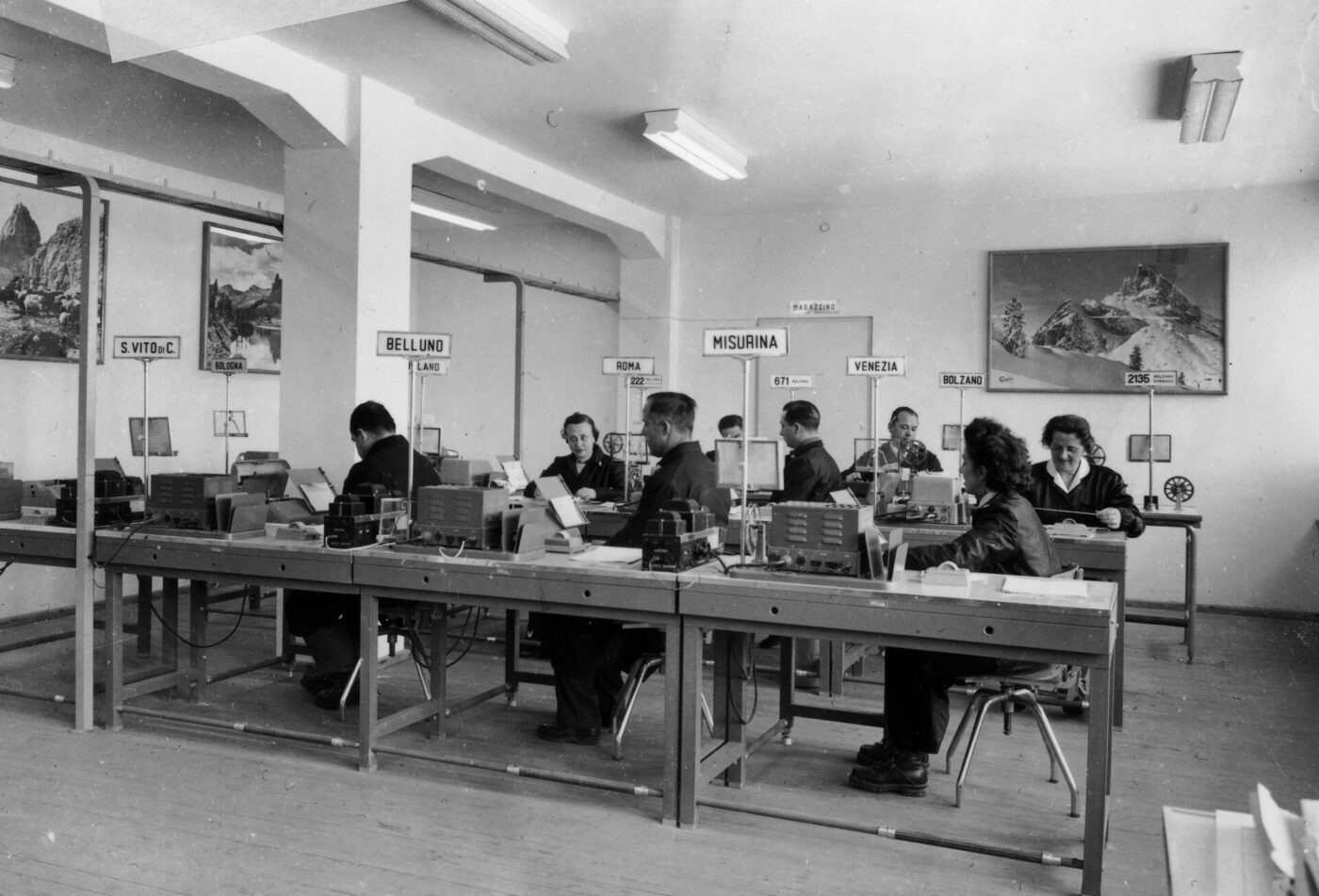 Una foto in bianco e nero mostra uomini e donne alle scrivanie di un ufficio con mappe, cartelli di toponimi alle pareti e attrezzature sui tavoli.