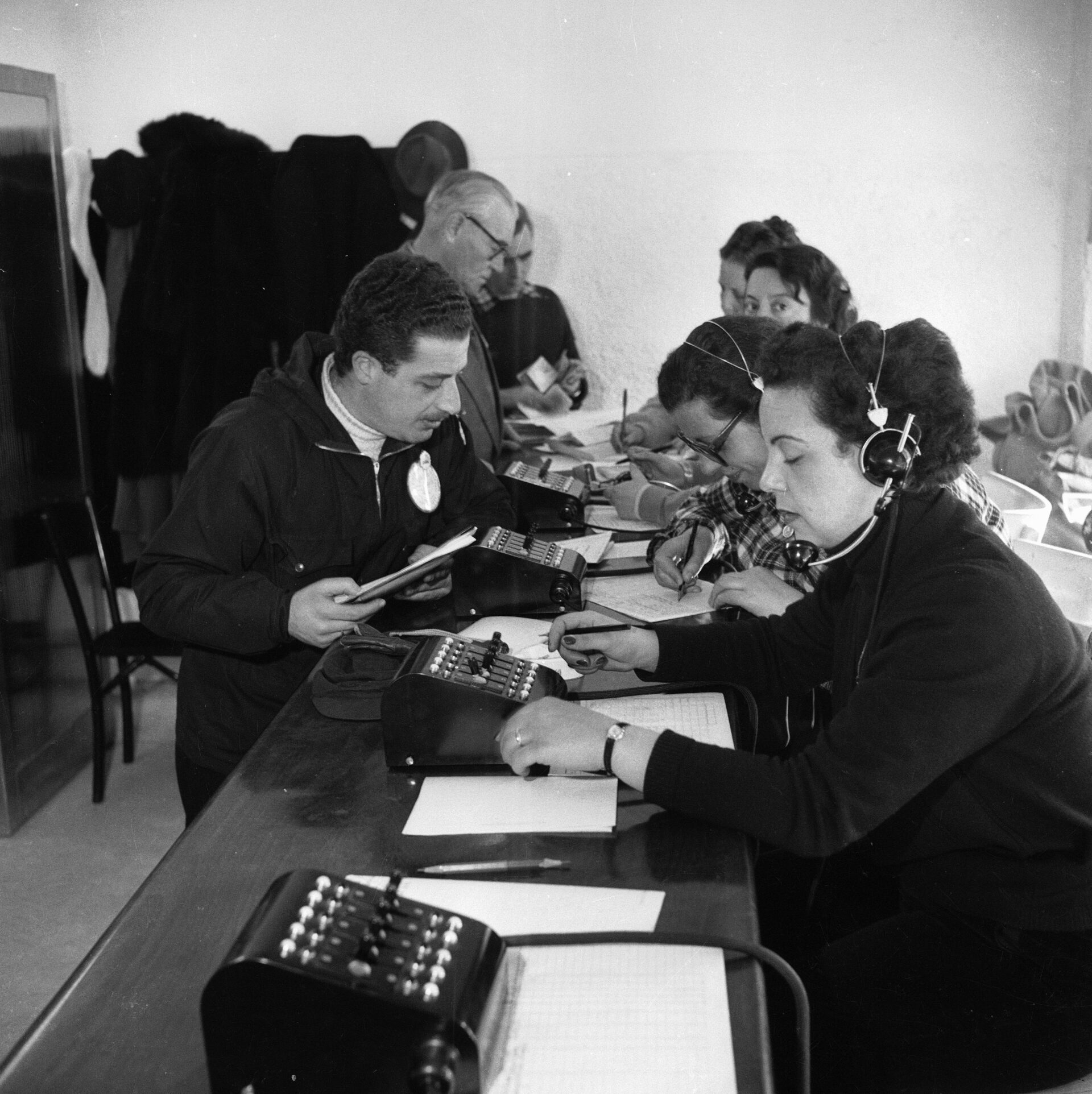 Persone che usano macchine da scrivere e calcolatrici a una lunga scrivania ricoperta di carte, alcune in piedi, una donna con le cuffie; ambiente d'ufficio.