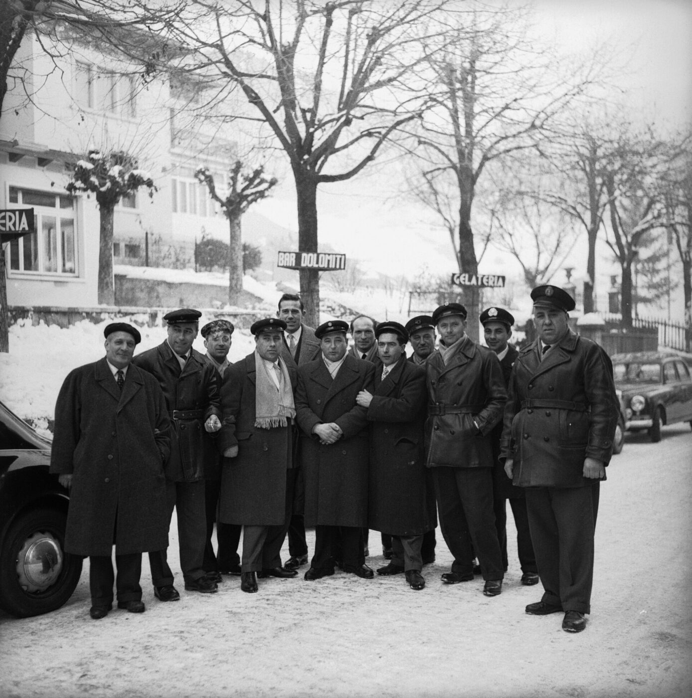 Un gruppo di uomini con cappotti e cappelli invernali si trova in una strada innevata; alle loro spalle sono visibili auto parcheggiate, alberi spogli e cartelli stradali.
