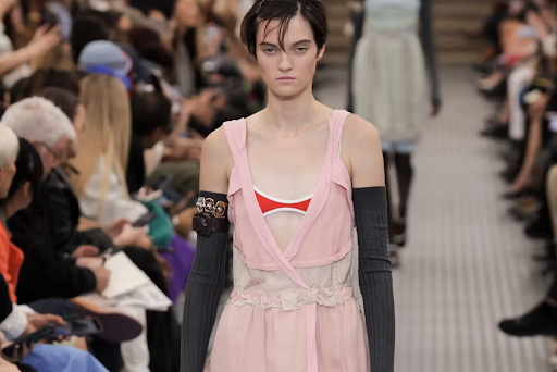 A model on the runway wears a pink dress, red bikini top, long gray arm warmers, upper arm accessories; audience lines the runway.