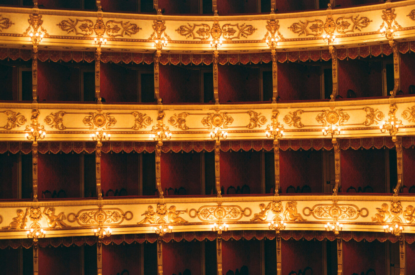 Rows of ornate, golden balconies with warm lights in a historic theater. Mostly empty red velvet seats and intricate decor create luxury.