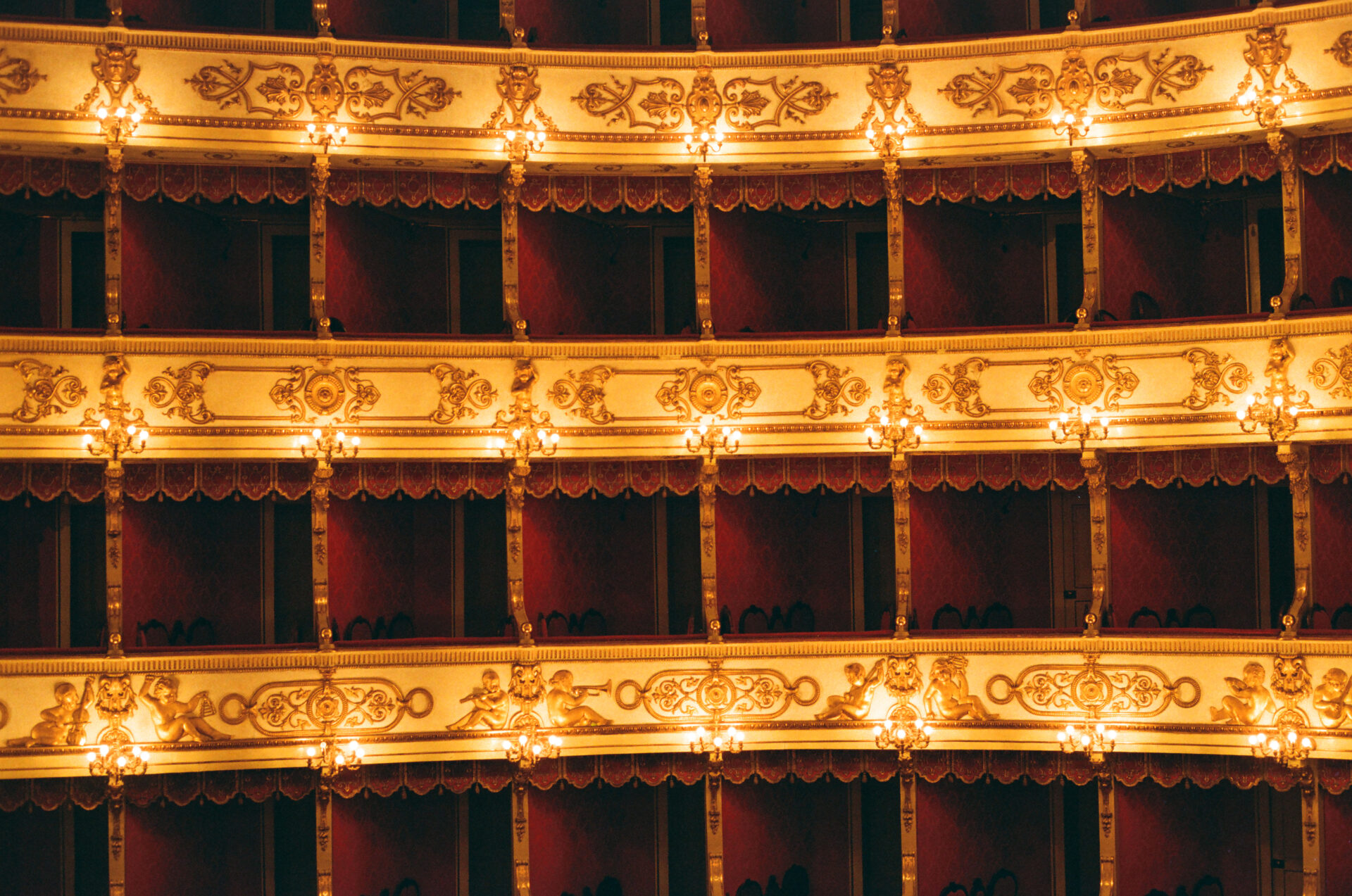 Rows of ornate, golden balconies with warm lights in a historic theater. Mostly empty red velvet seats and intricate decor create luxury.