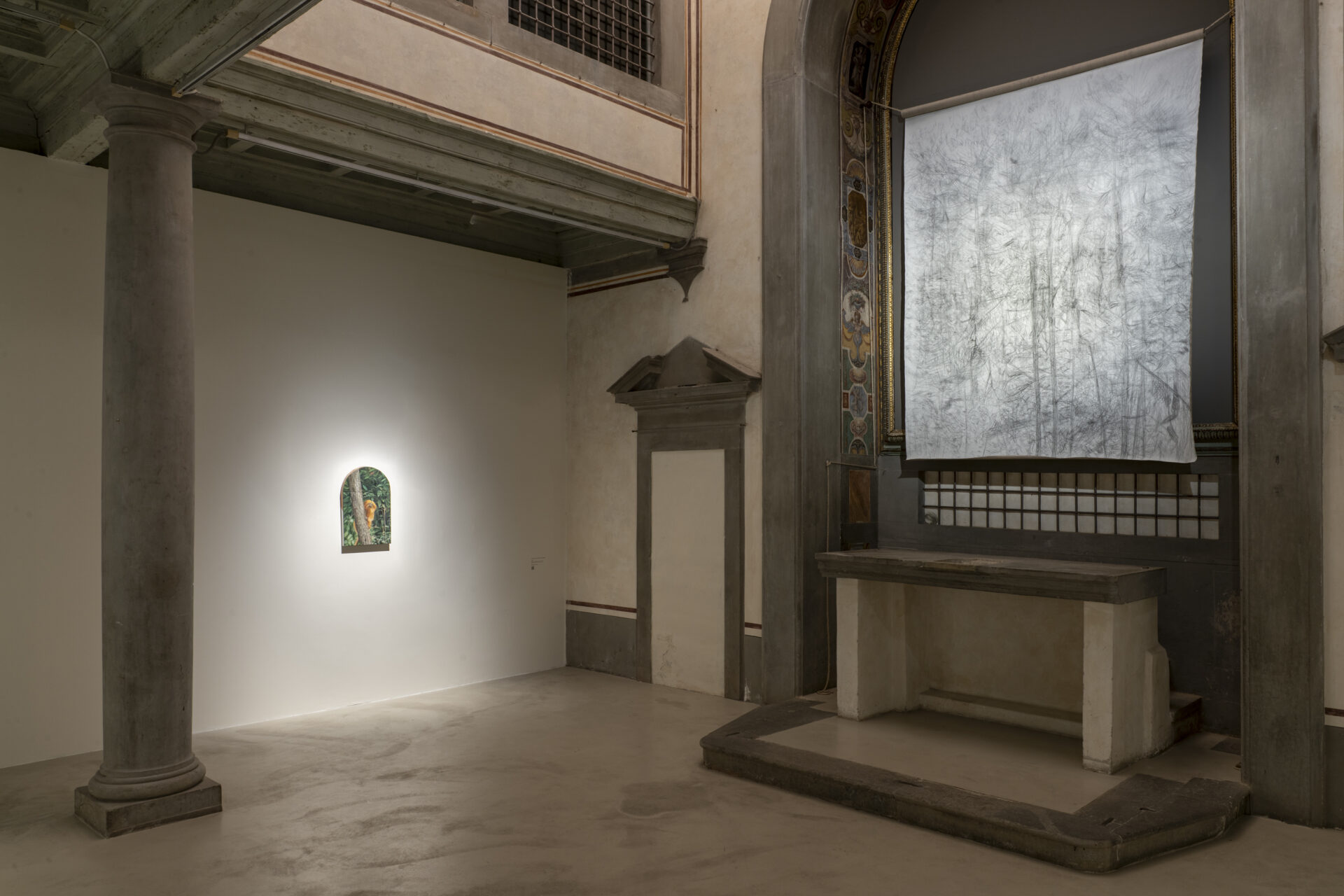 A gallery with classical columns, a small painting on a white wall, and textured silver art above an ornate altar. Soft lighting throughout.