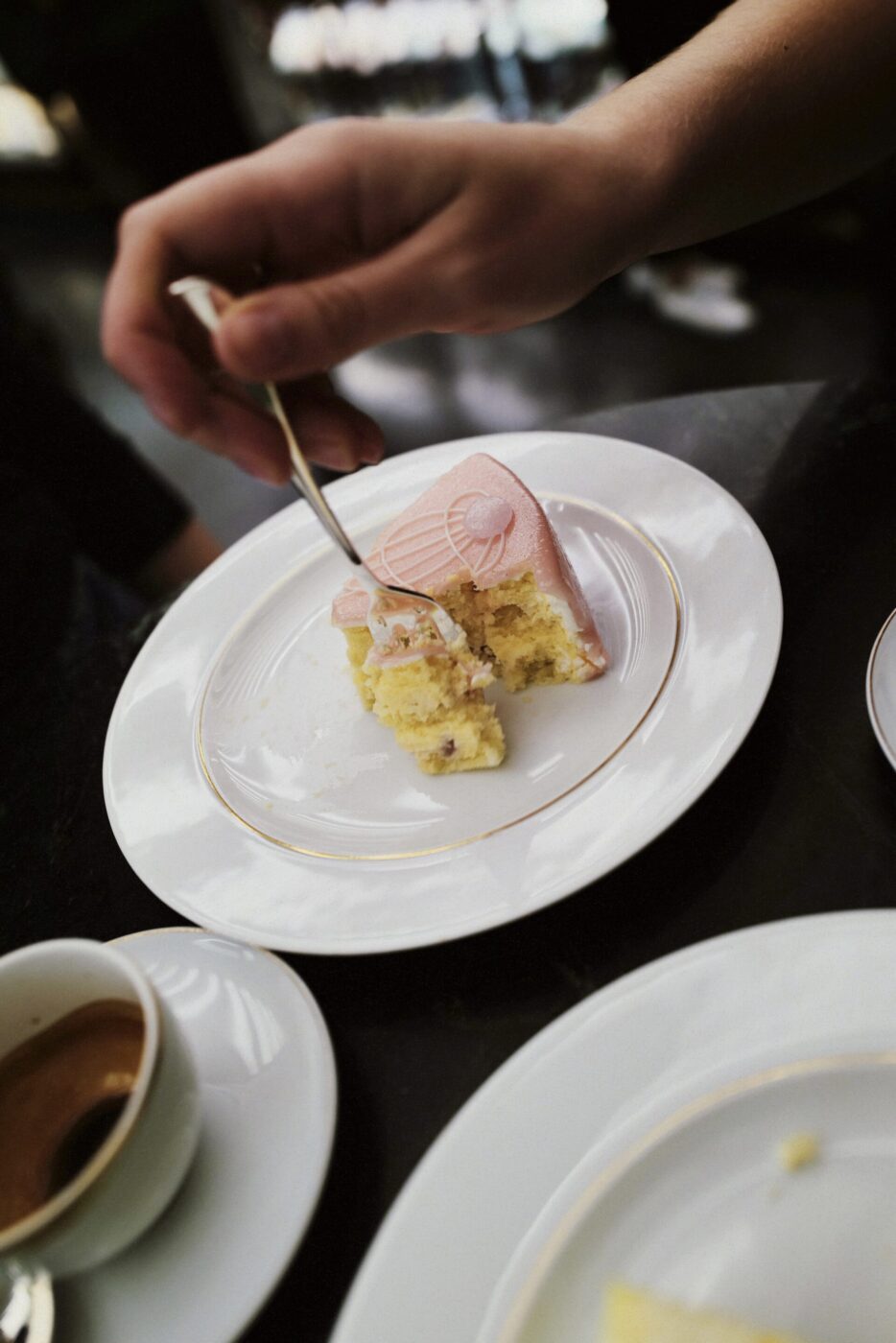 A hand with a fork slices into layered yellow cake with pink frosting; coffee and another plate sit on a dark table nearby.