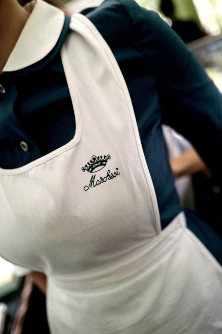 A person in a dark blue collared shirt and white Marchesi apron with crown embroidery; background softly blurred.