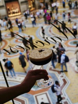 A hand holds a foamy espresso martini, seen from above through a window; busy, colorful patterned floor and people below.