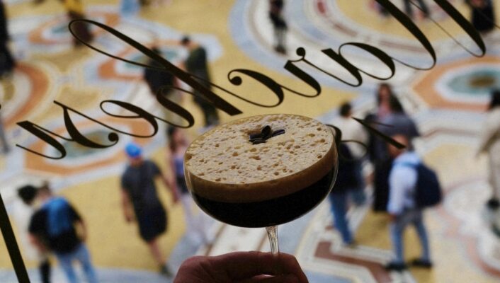 A hand holds a foamy espresso martini, seen from above through a window; busy, colorful patterned floor and people below.