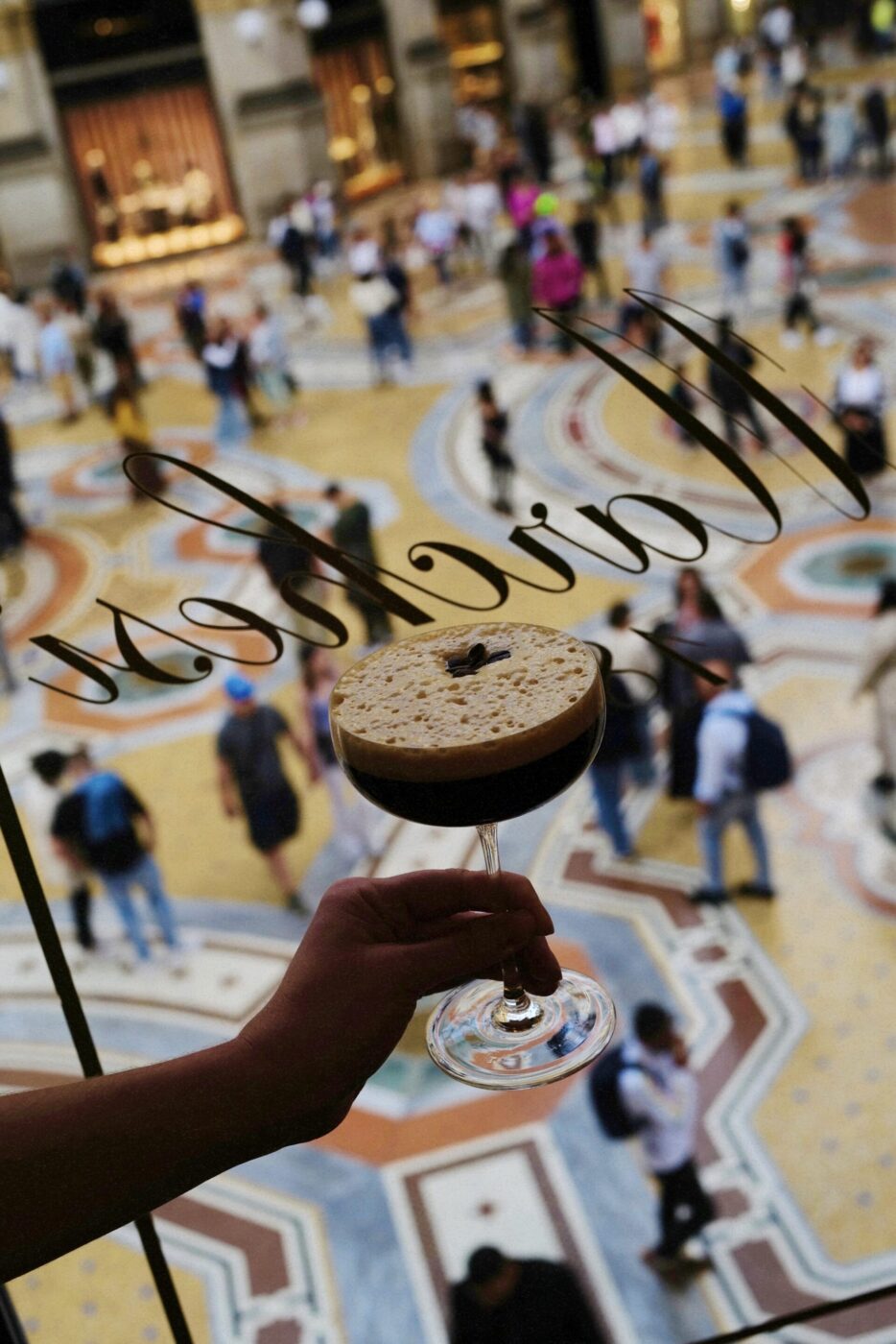 A hand holds a foamy espresso martini, seen from above through a window; busy, colorful patterned floor and people below.